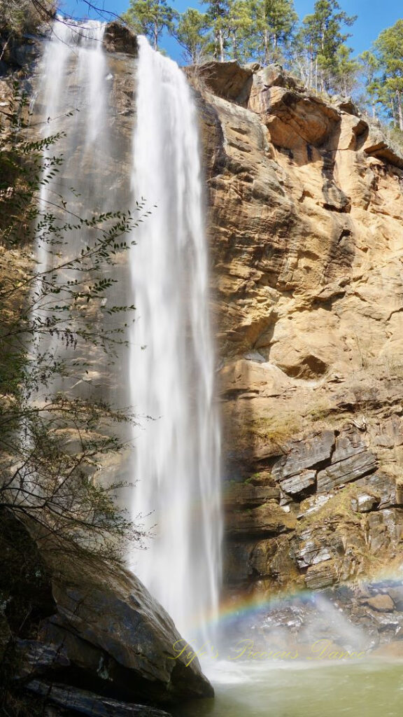 An 186 foot waterfall spilling over a rock face into a pool of water at Toccoa Falls. A rainbow is reflecting in the mist.