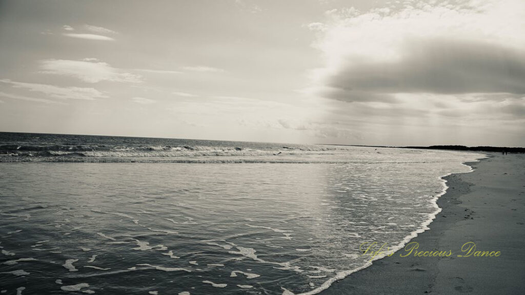 Black and white seascape view of Huntington Beach. Clouds above and waves crashing against the shore.