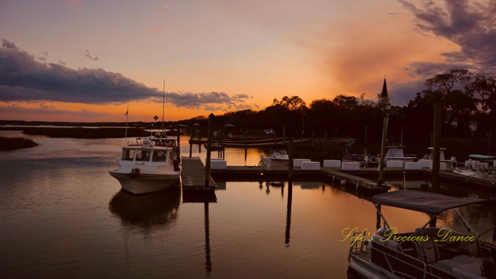 Boats resting along a dock and reflecting in the water at Murrells Inlet. The sun is setting in the background.