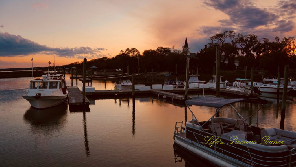 Boats resting along a dock and reflecting in the water at Murrells Inlet. The sun is setting in the background.