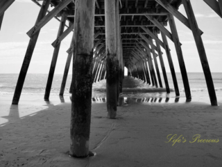 Black and white view from under the pier as ocean waves crash against the beach.