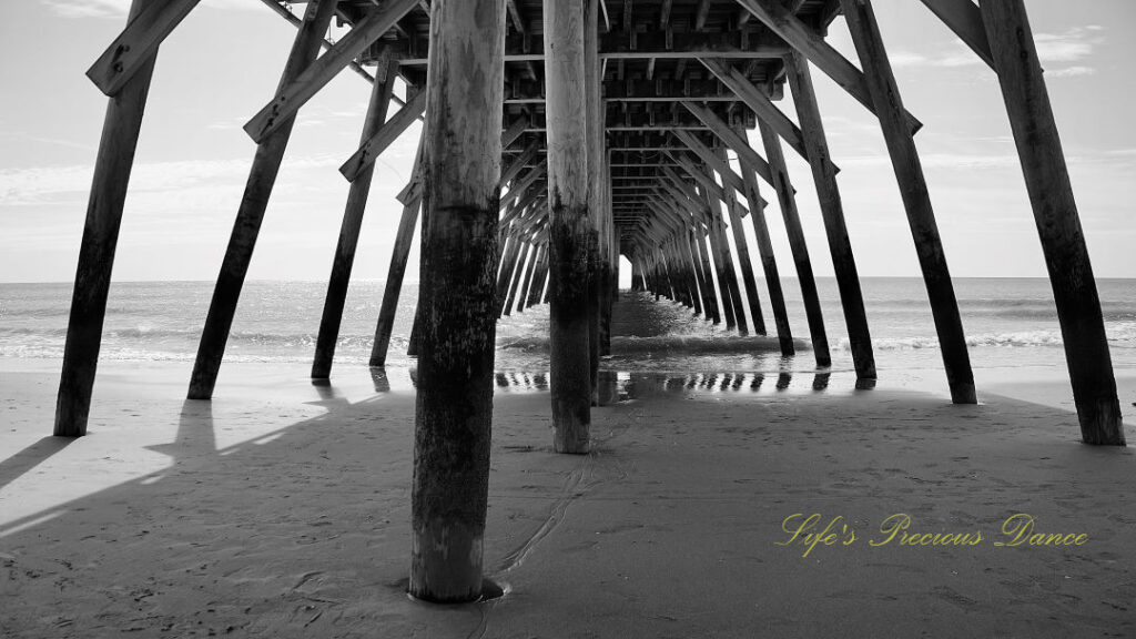 Black and white view from under the pier as ocean waves crash against the beach.