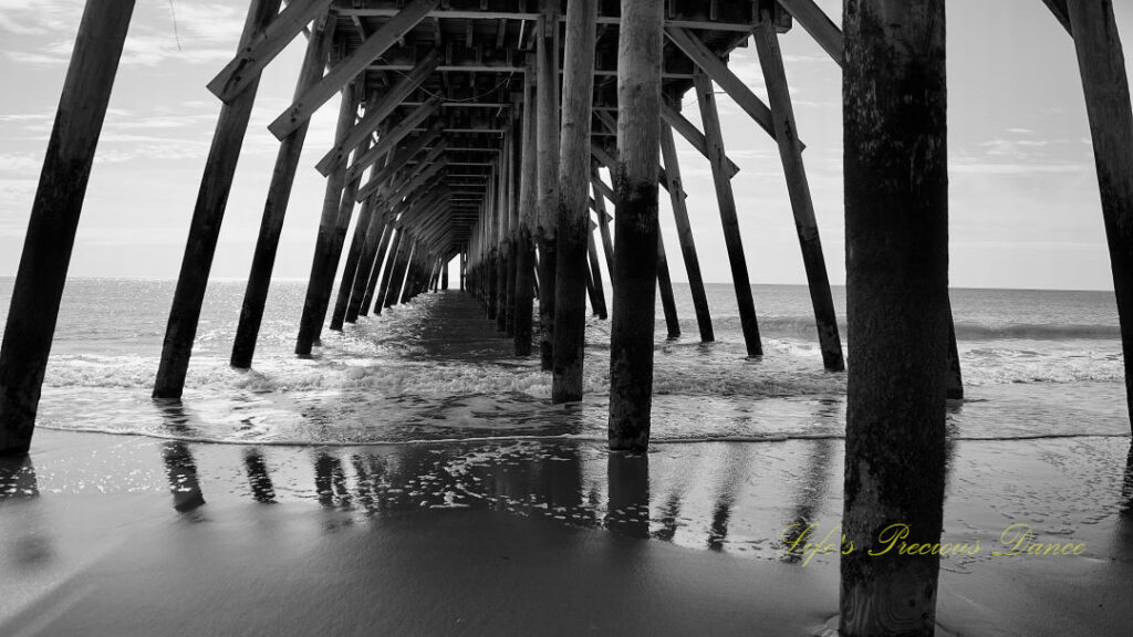 Black and white view from under the pier as ocean waves crash against the beach.