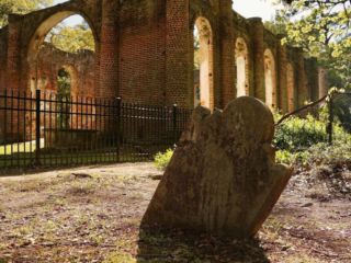 Close up of an old slanted tombstone, sinking in the ground. The Old Sheldon Church ruins in the background.