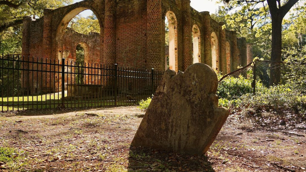 Close up of an old slanted tombstone, sinking in the ground. The Old Sheldon Church ruins in the background.