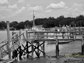 Black and white of a pier at Jeremy Creek landing. Passing clouds overhead and shrimp boats in the background.