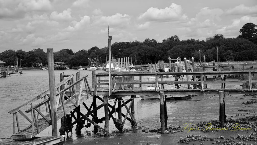 Black and white of a pier at Jeremy Creek landing. Passing clouds overhead and shrimp boats in the background.