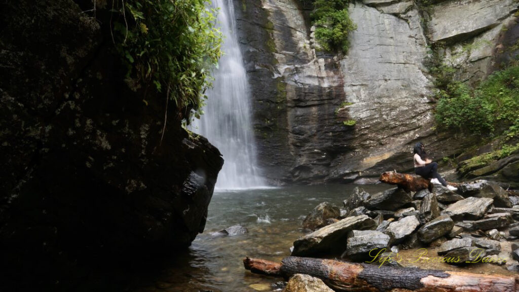 Looking Glass Falls spilling down a rockface into the creek below. A large boulder in the foreground to the left partially blocks its view. A young lady sits on a downed tree to the right.