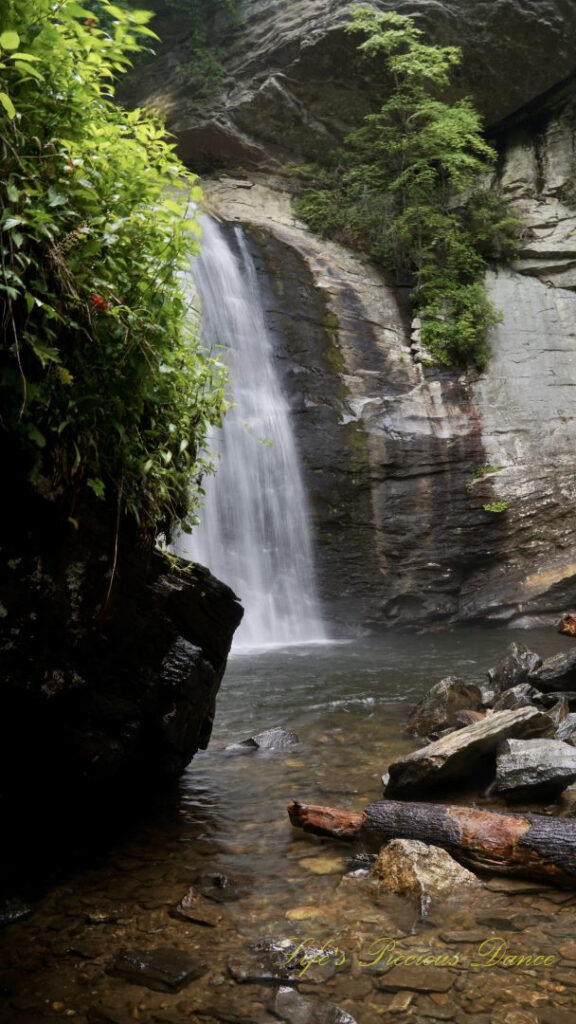 Looking Glass Falls spilling down a rockface into the creek below. A large boulder in the foreground to the left partially blocks its view.