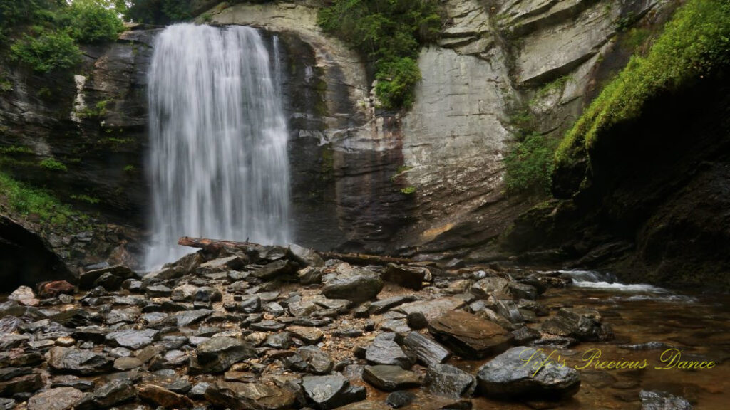 Looking Glass Falls spilling into the awaiting creek below. Scattered rocks and a downed tree in the foreground.