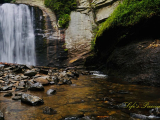 Looking Glass Falls spilling into the awaiting creek below. Scattered rocks and a downed tree in the foreground.