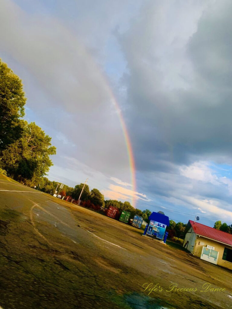 Rainbow stretching up above a parking lot with colorful shipping containers. Clouds in the background.