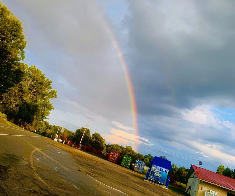 Rainbow stretching up above a parking lot with colorful shipping containers. Clouds in the background.