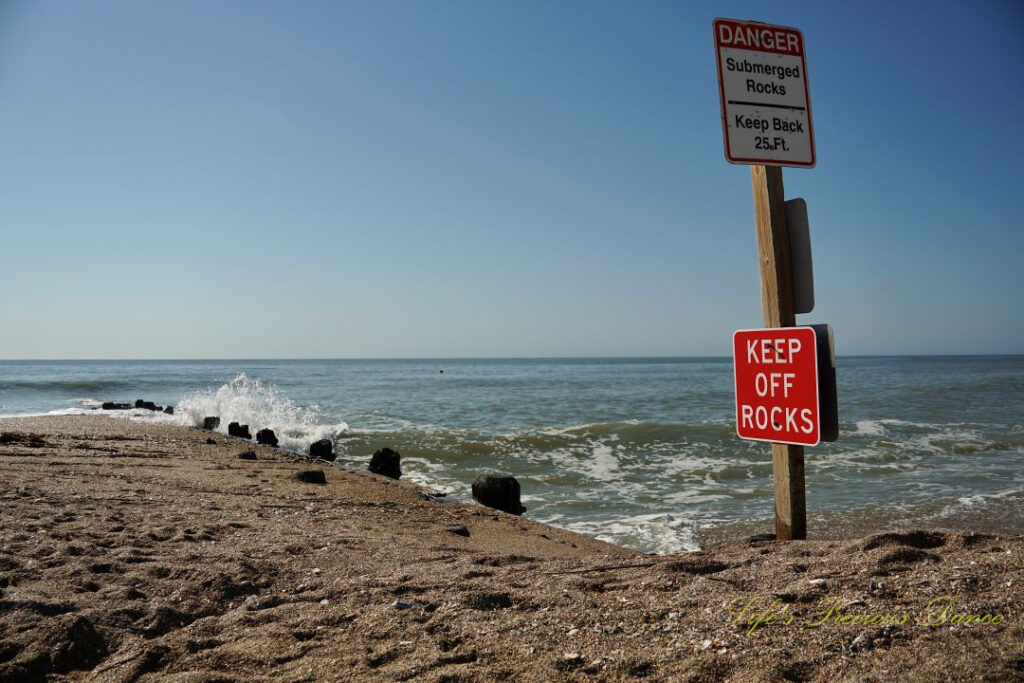 Warning sign to Keep Off Rocks at Edisto Beach State Park. Atlantic Ocean in the background and waves crashing into a barrier.