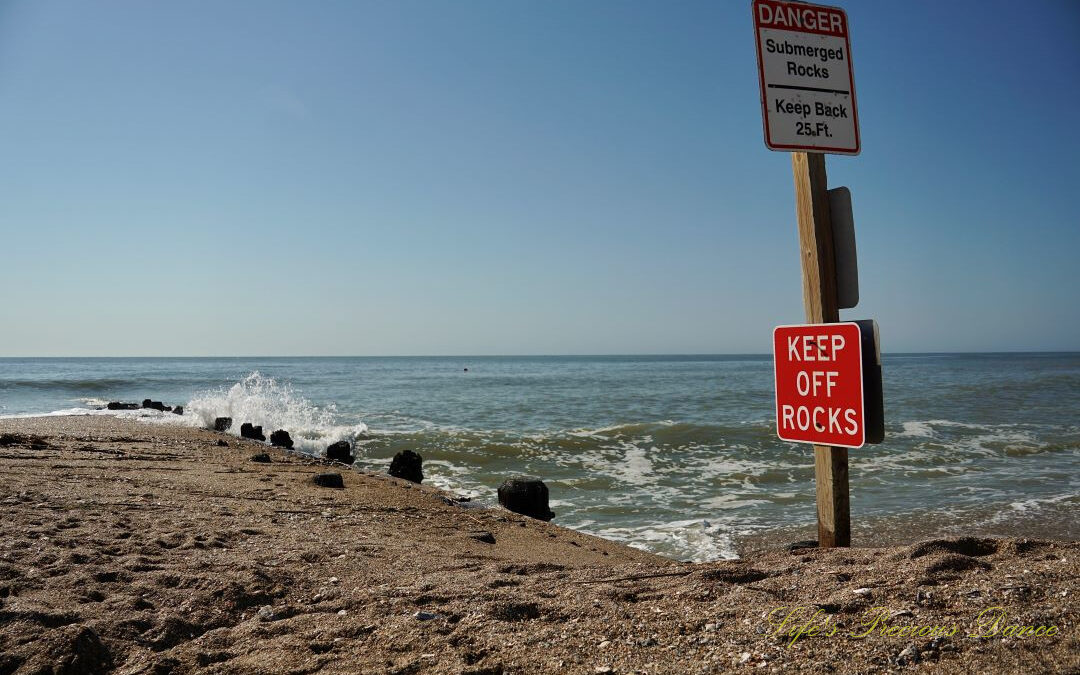 Warning sign to Keep Off Rocks at Edisto Beach State Park. Atlantic Ocean in the background and waves crashing into a barrier.