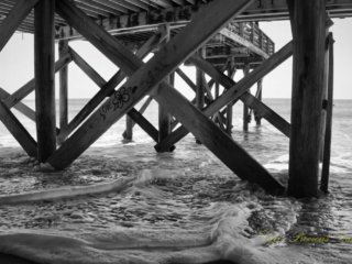 Black and white underside view of the pier at Edisto Beach. Graffiti on a support beam with a flower and the word Love. Frothy waves covering the sand.