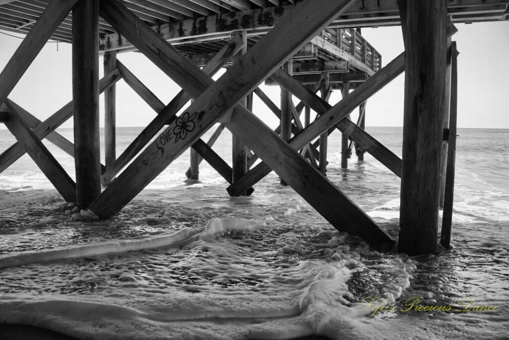 Black and white underside view of the pier at Edisto Beach. Graffiti on a support beam with a flower and the word Love. Frothy waves covering the sand.