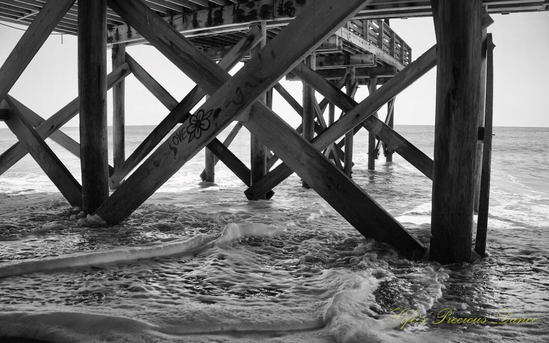 Black and white underside view of the pier at Edisto Beach. Graffiti on a support beam with a flower and the word Love. Frothy waves covering the sand.