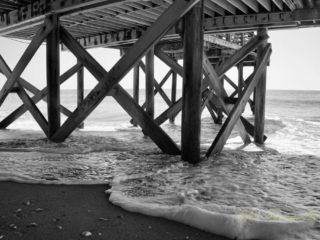 Black and white underside of the pier at Edisto Beach. Seashells in the sand and frothy waves rolling in.
