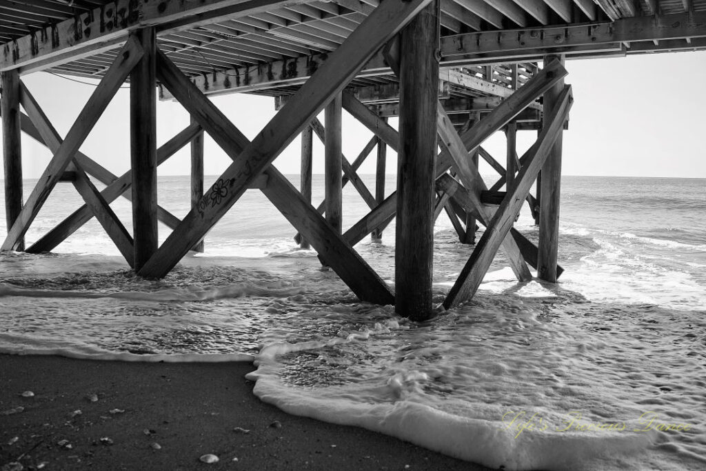 Black and white underside of the pier at Edisto Beach. Seashells in the sand and frothy waves rolling in.