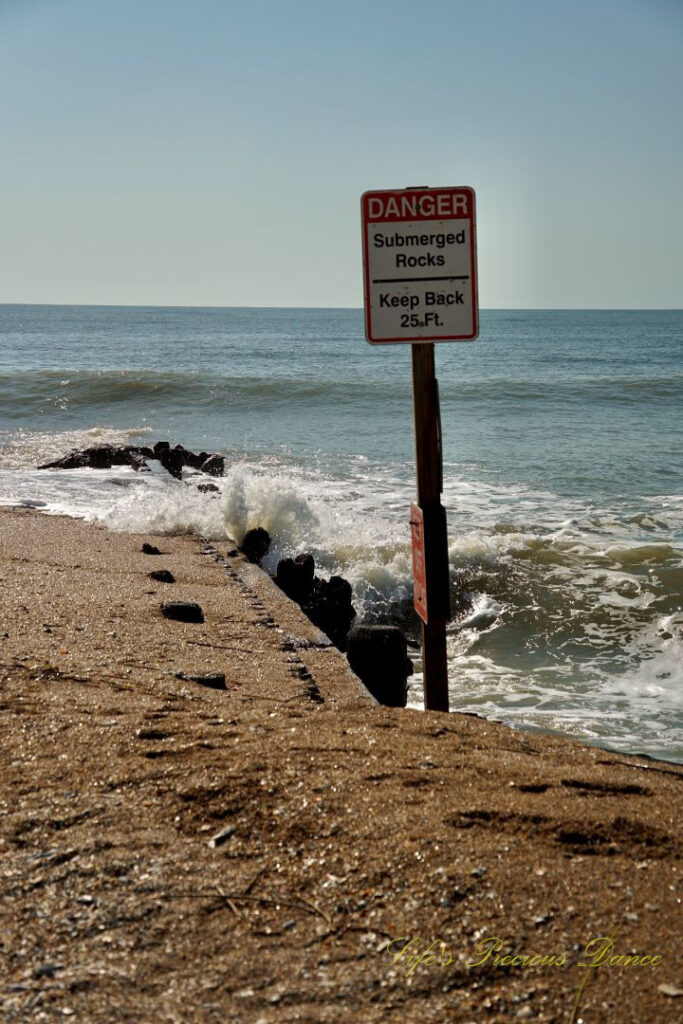 Warning sign stating the Danger of Submerged Rocks at Edisto Beach State Park. Waves crashing into a barrier in the background.