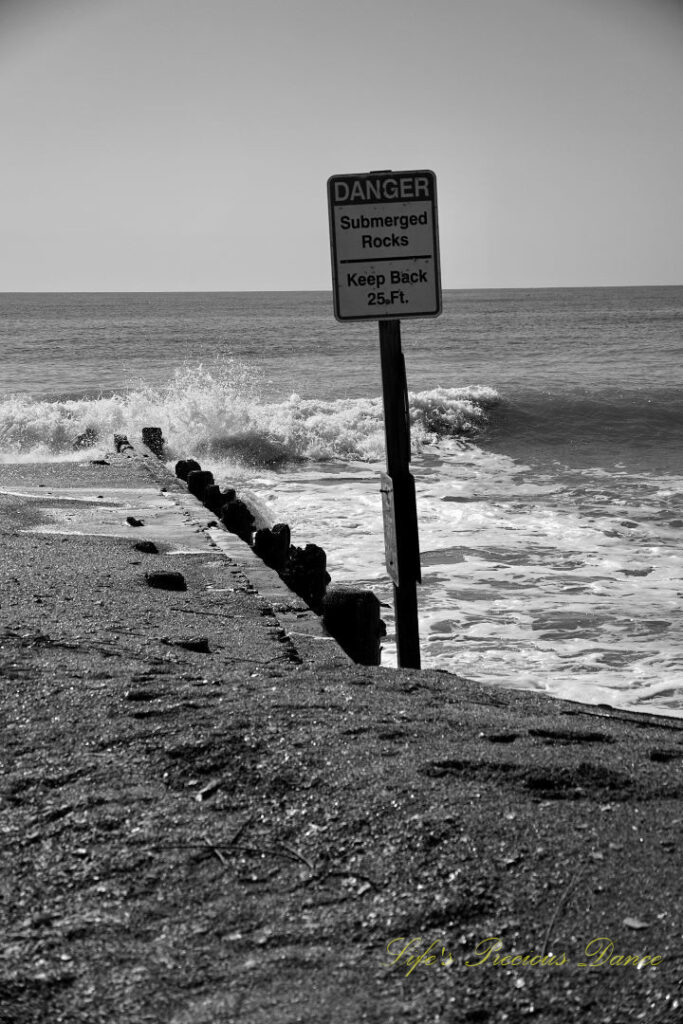 Black and white warning sign stating the Danger of Submerged Rocks at Edisto Beach State Park. Waves crashing into a barrier in the background.