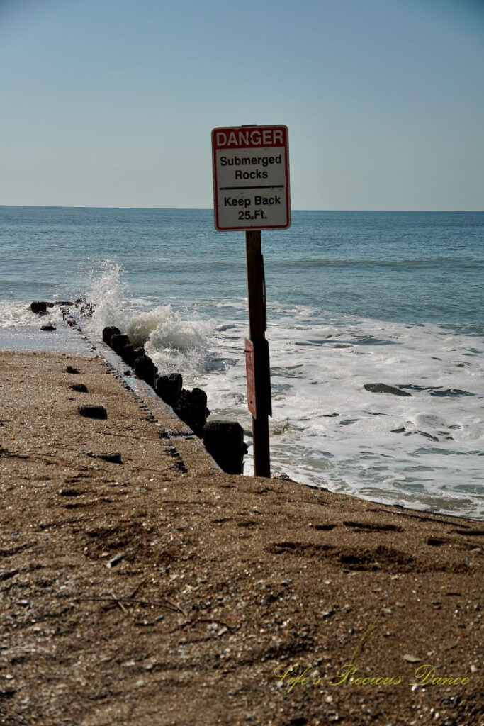 Warning sign stating the Danger of Submerged Rocks at Edisto Beach State Park. Waves crashing into a barrier in the background.