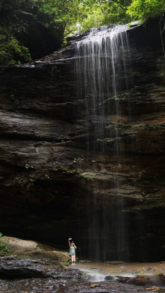 Moore Cove Falls spilling over the rockledge into the mountain stream. A woman taking a selfie at the base.