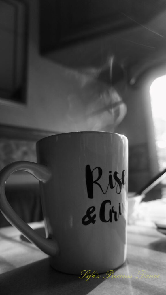 Black and white of a coffee mug on a table with steam rising from the top. The sun&#039;s rays streaking through a window.