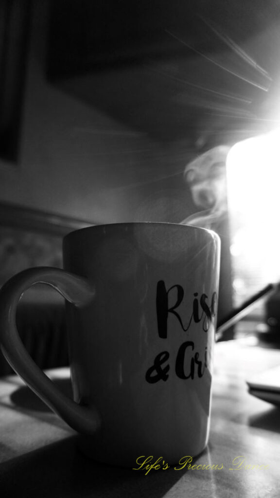 Black and white of a coffee mug on a table with steam rising from the top. The sun&#039;s rays streaking through a window.