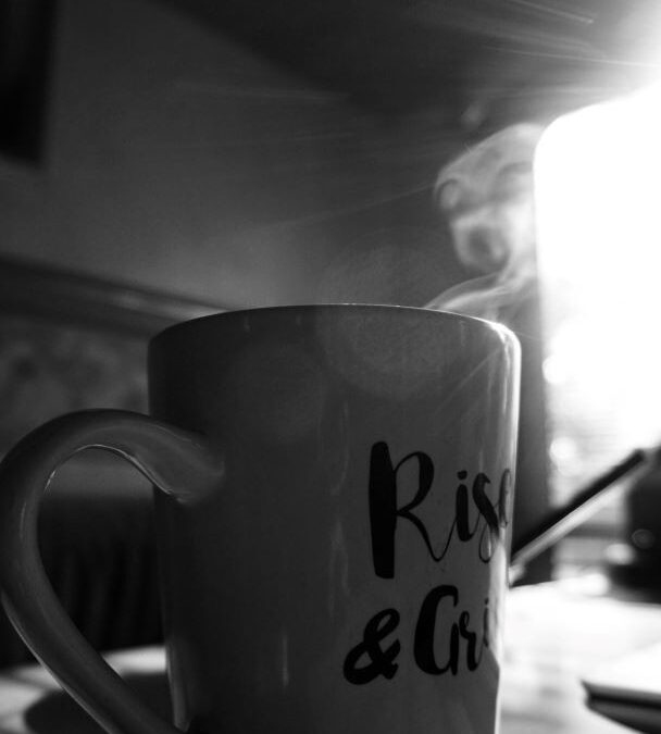 Black and white of a coffee mug on a table with steam rising from the top. The sun's rays streaking through a window.