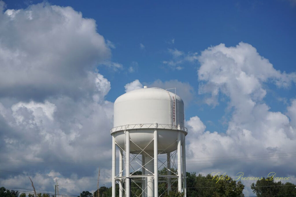 Top section of a water tower. Passing fluffy clouds and trees in the background.