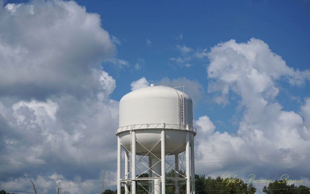 Top section of a water tower. Passing fluffy clouds and trees in the background.