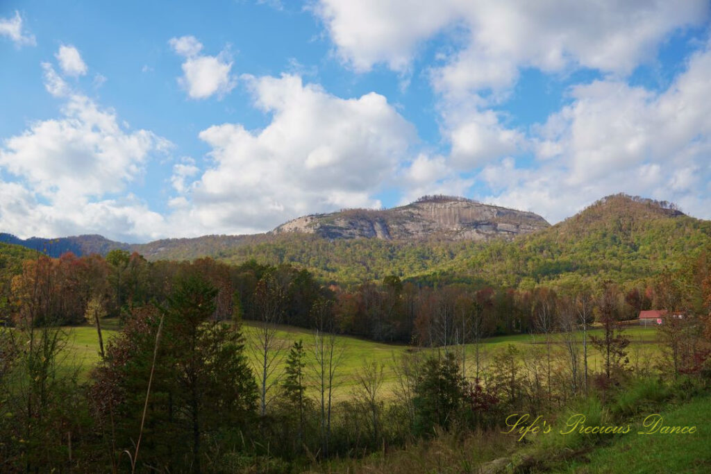 Landscape view of Table Rock and the surrounding mountain range from Grant Meadow Overlook. Passing fluffy clouds overhead. A small ranch house can be seen to the right.