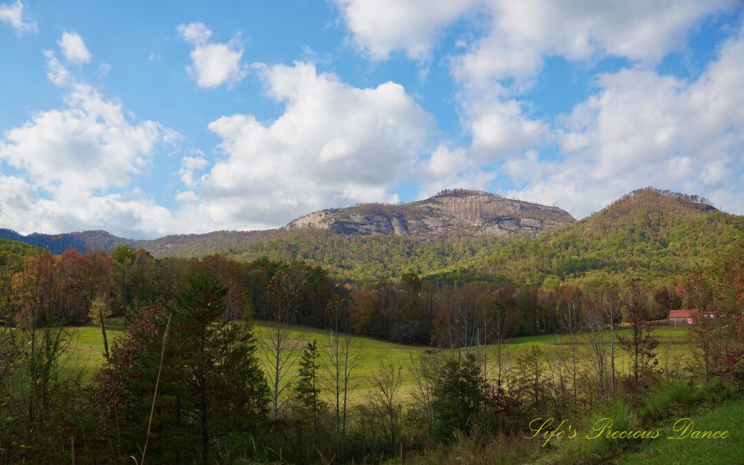 Landscape view of Table Rock and the surrounding mountain range from Grant Meadow Overlook. Passing fluffy clouds overhead. A small ranch house can be seen to the right.