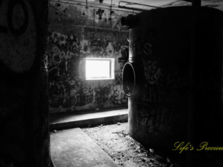 Black and white of the graffiti covered pump room, located below the water level at abandoned Springs pool. A viewing window can be seen on a backwall.