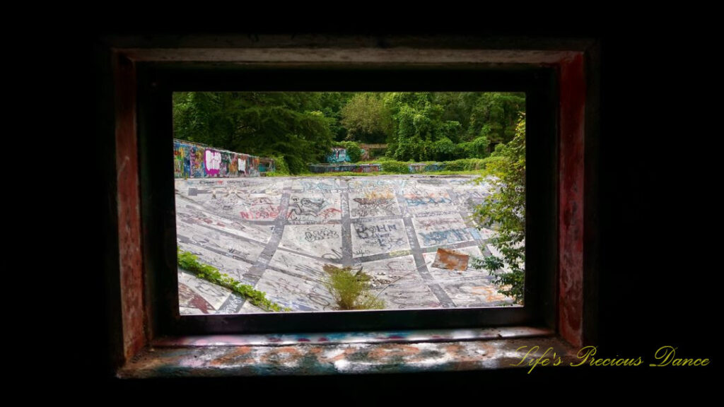 Close up of a window, surrounded by graffiti, looking outward at the abandoned Springs Recreation pool.