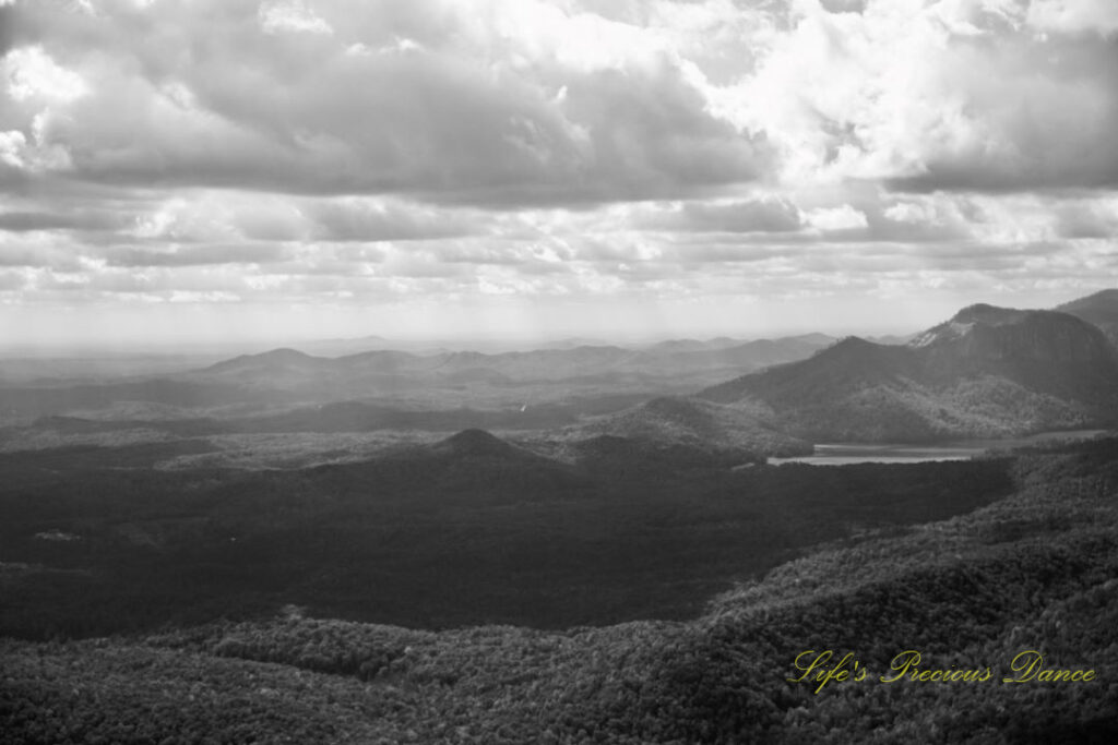 Black and white landscape view from Caesars Head Overlook. Passing clouds overhead. A lake can be seen below the distant mountain range.