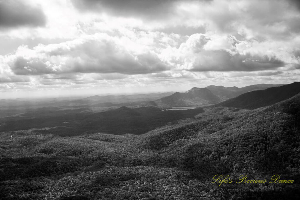 Black and white landscape view from Caesars Head Overlook. Passing clouds overhead. A lake can be seen below the distant mountain range.