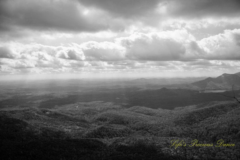 Black and white landscape view from Caesars Head Overlook. Passing clouds overhead. A lake can be seen below the distant mountain range.