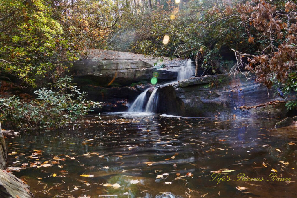Poe Creek Falls spilling over the rocks into the creek as the sun&#039;s rays shine down. Leaves floating on the waters surface and colorful trees surrounding the scene.