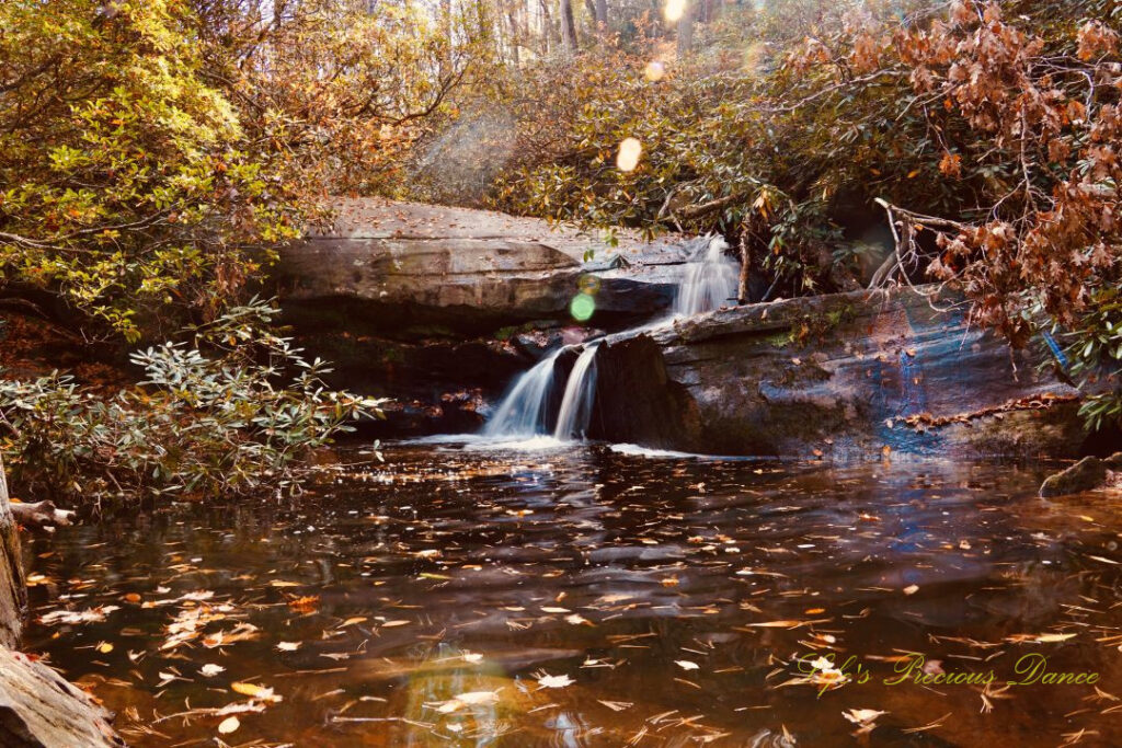 Poe Creek Falls spilling over the rocks into the creek as the sun&#039;s rays shine down. Leaves floating on the waters surface and colorful trees surrounding the scene.