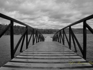 Black and white ground level view looking straight down a pier leading out into Lake Jocassee. Passing clouds overhead and a mountain range in the background.