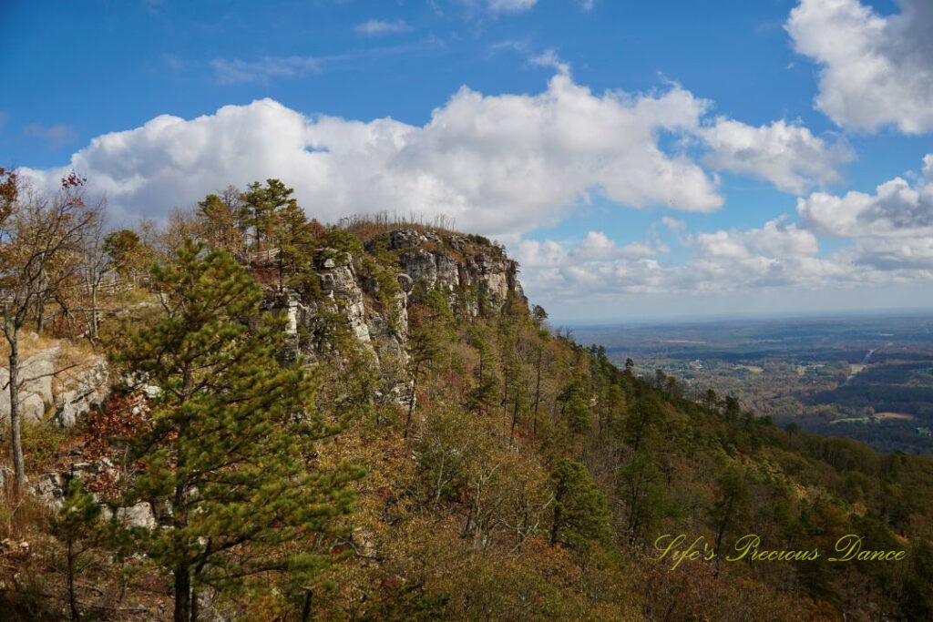 Looking at the west face of Pilot Mountain from a viewing deck. Passing clouds against a blue sky in the background and colorful trees in the valley below.