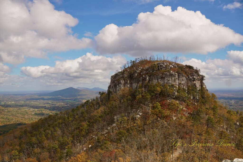 View of the north face of Pilot Mountain surrounded by colorful trees. Passing clouds overhead and distant mountain peaks in the background.