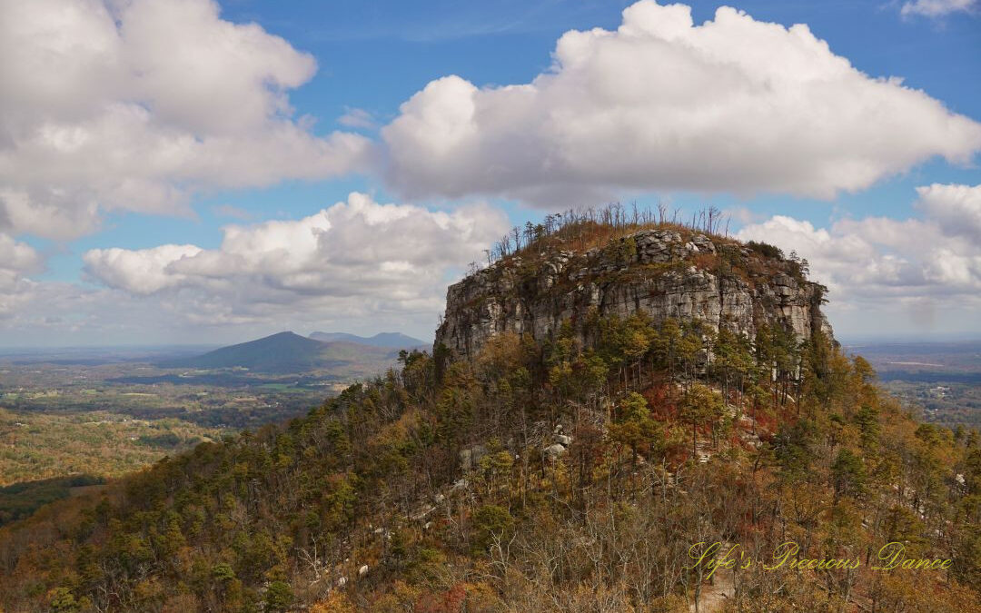 View of the north face of Pilot Mountain surrounded by colorful trees. Passing clouds overhead and distant mountain peaks in the background.