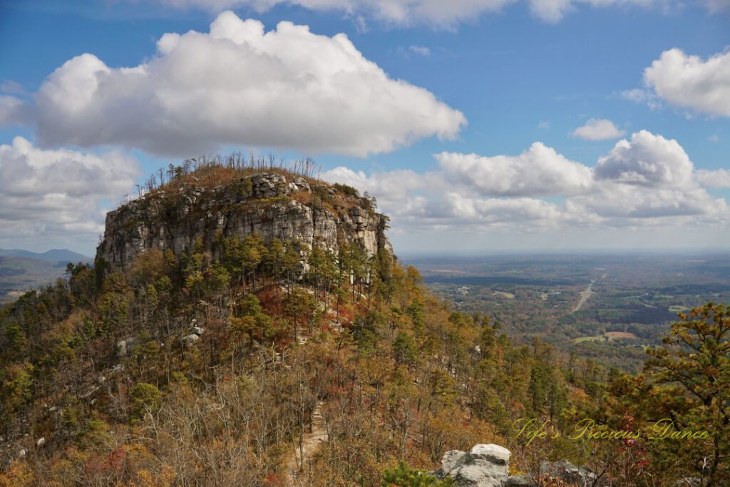 View of the north face of Pilot Mountain surrounded by colorful trees. Passing clouds overhead and distant mountain peaks in the background.