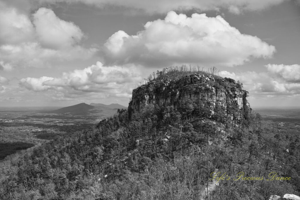 Black and white view of the north face of Pilot Mountain surrounded by colorful trees. Passing clouds overhead and distant mountain peaks in the background.
