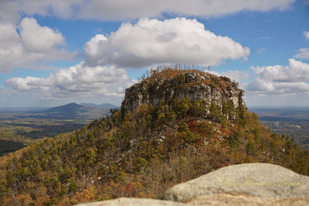 View of the north face of Pilot Mountain surrounded by colorful trees. Passing clouds overhead and distant mountain peaks in the background.