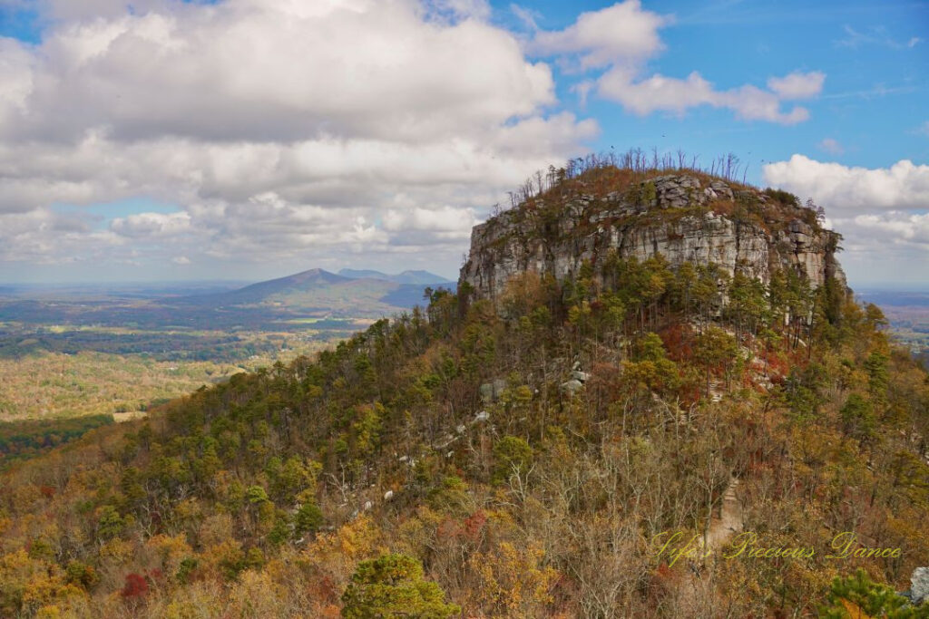 View of the north face of Pilot Mountain surrounded by colorful trees. Passing clouds overhead and distant mountain peaks in the background.